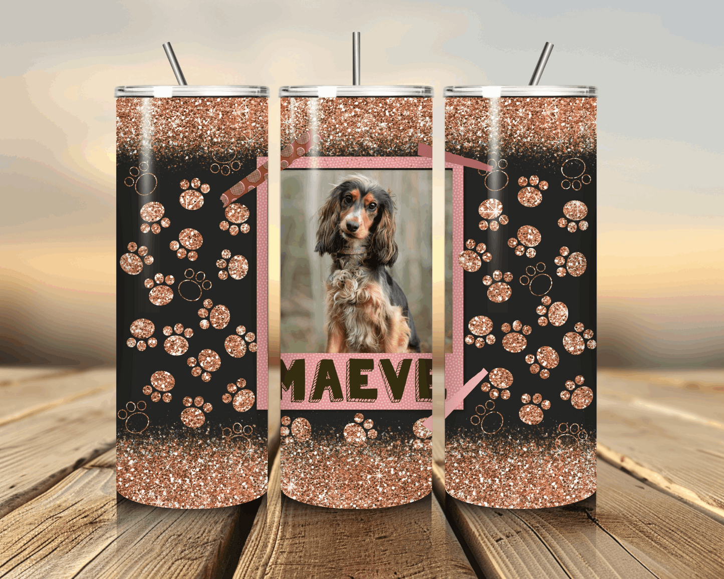 Three glittery tumblers with paw patterns and a photo of a dog named Maeve on a wooden surface.