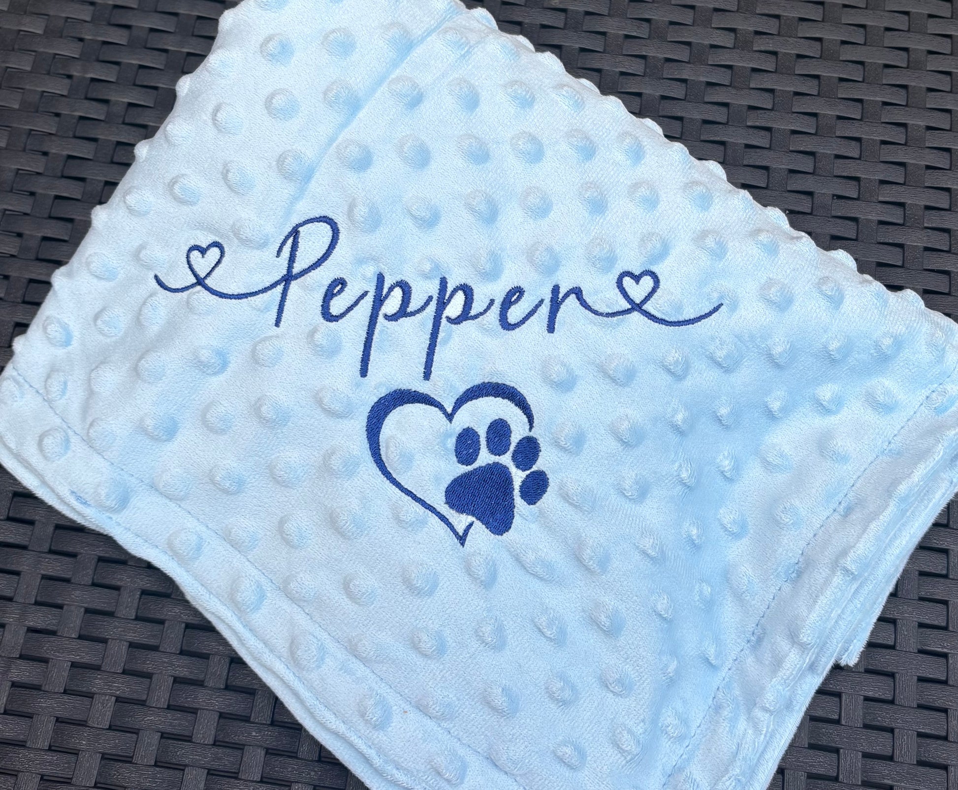 Light blue textured blanket with 'Pepper' and a paw print design on a dark woven surface
