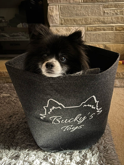 Dog peeking out from a black bag with 'Bucky's Toys' branding against a brick wall background.