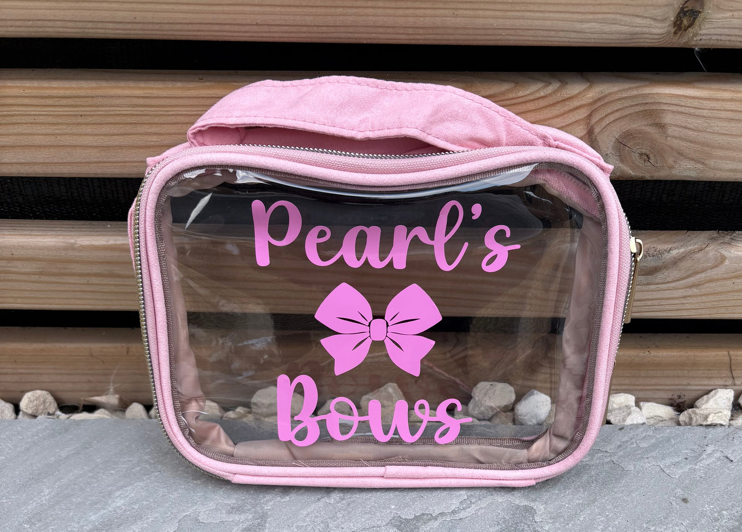 Clear vanity bag with pink zipper and 'Pearl's Bows' text and bow design on a wooden background