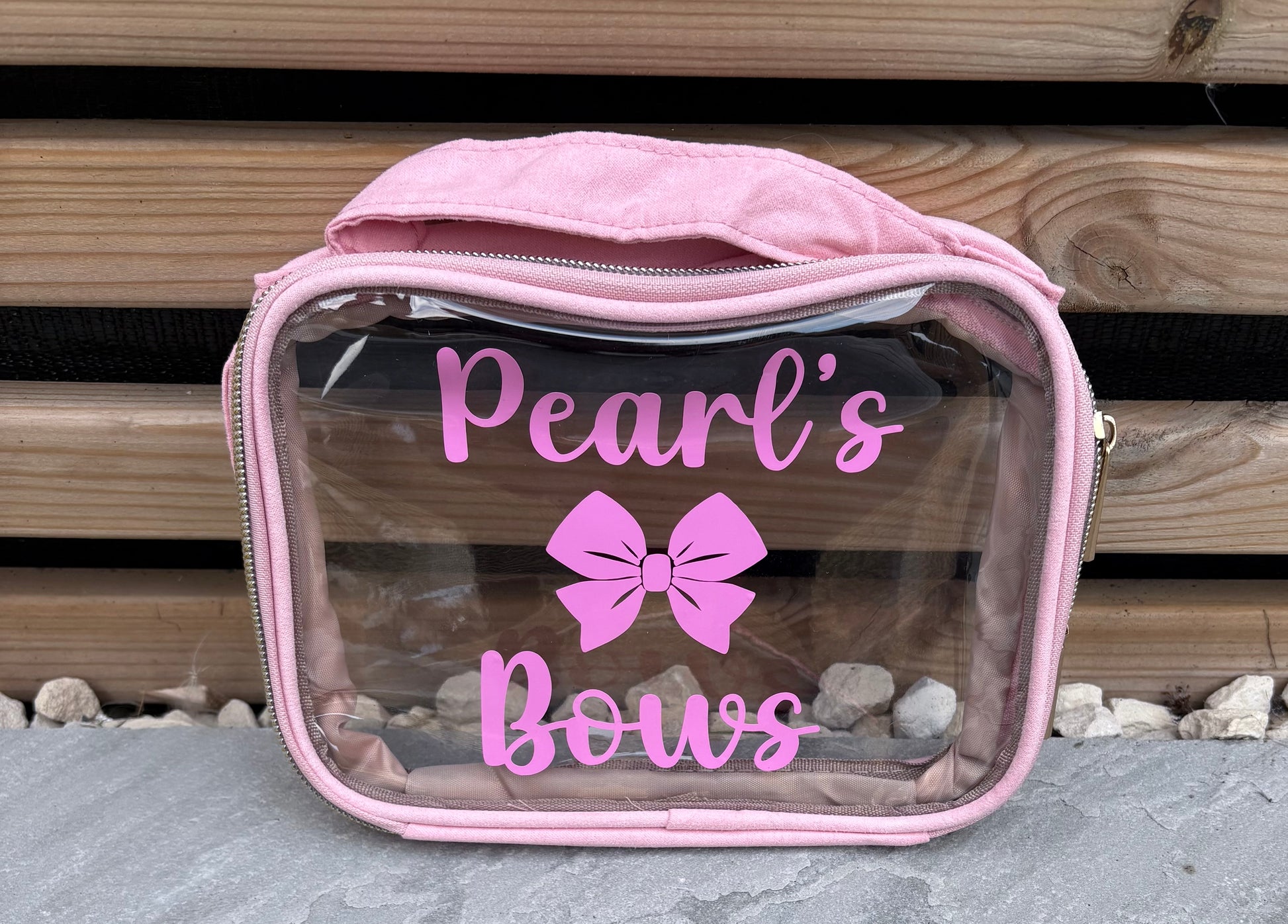 Clear vanity bag with pink zipper and 'Pearl's Bows' text and bow design on a wooden background