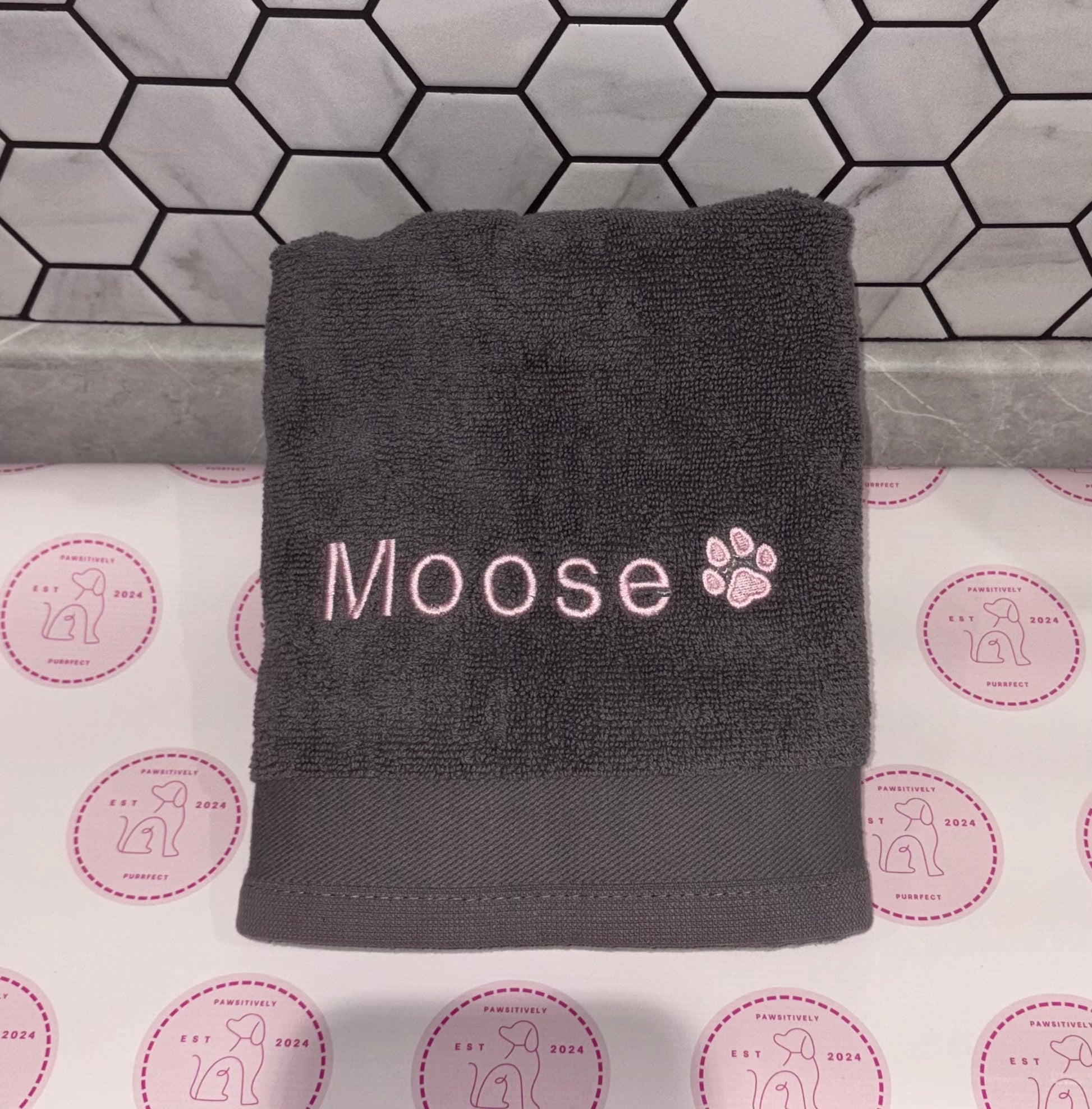Gray towel with 'Moose' and paw print on a tiled bathroom floor