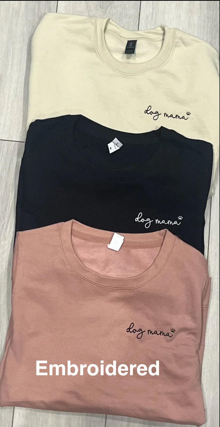 Three folded sweatshirts in beige, black, and pink with 'dog mama' embroidery on a wooden surface.