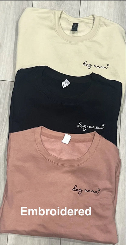 Three folded sweatshirts in beige, black, and pink with 'dog mama' embroidery on a wooden surface.