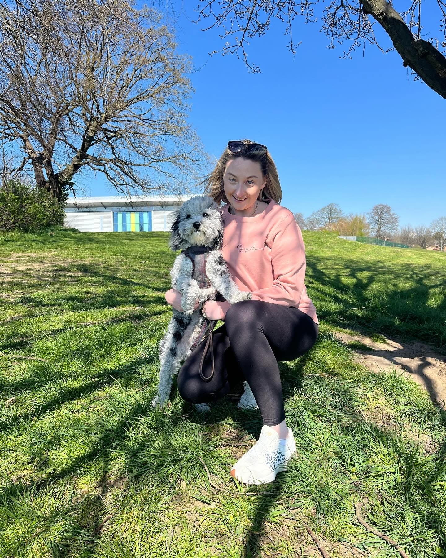 Woman sitting on grass with a dog, wearing a pink sweater and sunglasses.