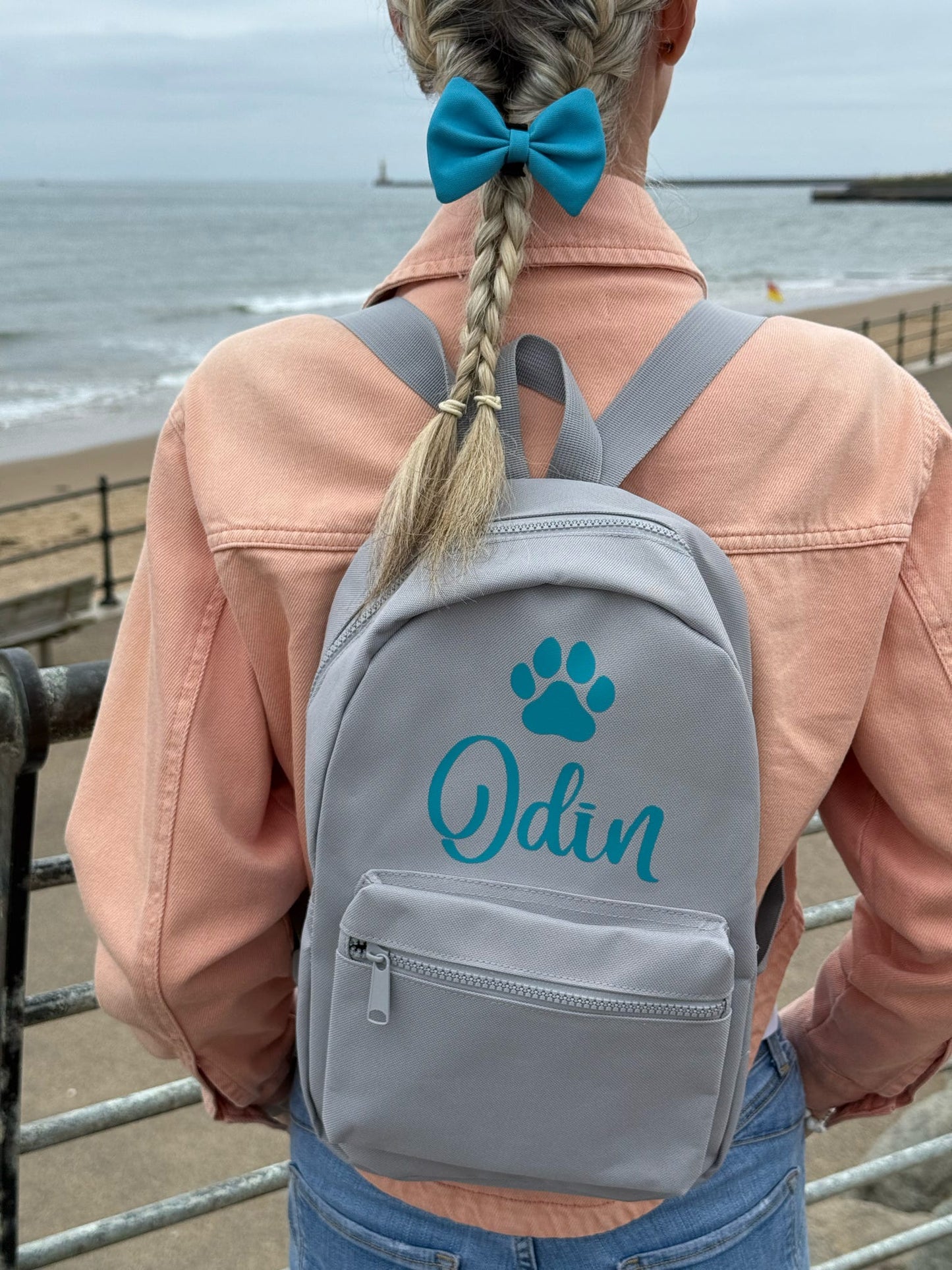 Person wearing a gray backpack with 'Odin' and a paw print design, standing by the sea.