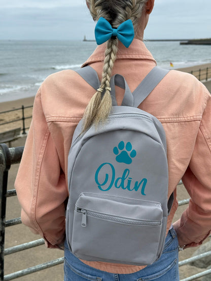 Person wearing a gray backpack with 'Odin' and a paw print design, standing by the sea.