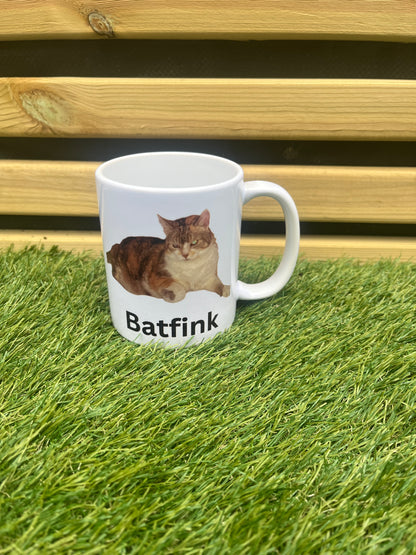 White mug with a cat design and 'Batfink' text on grass and wooden surface