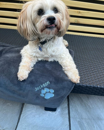 Small white dog sitting on a gray blanket with 'Jackson' and a paw print on it, on a wooden deck.